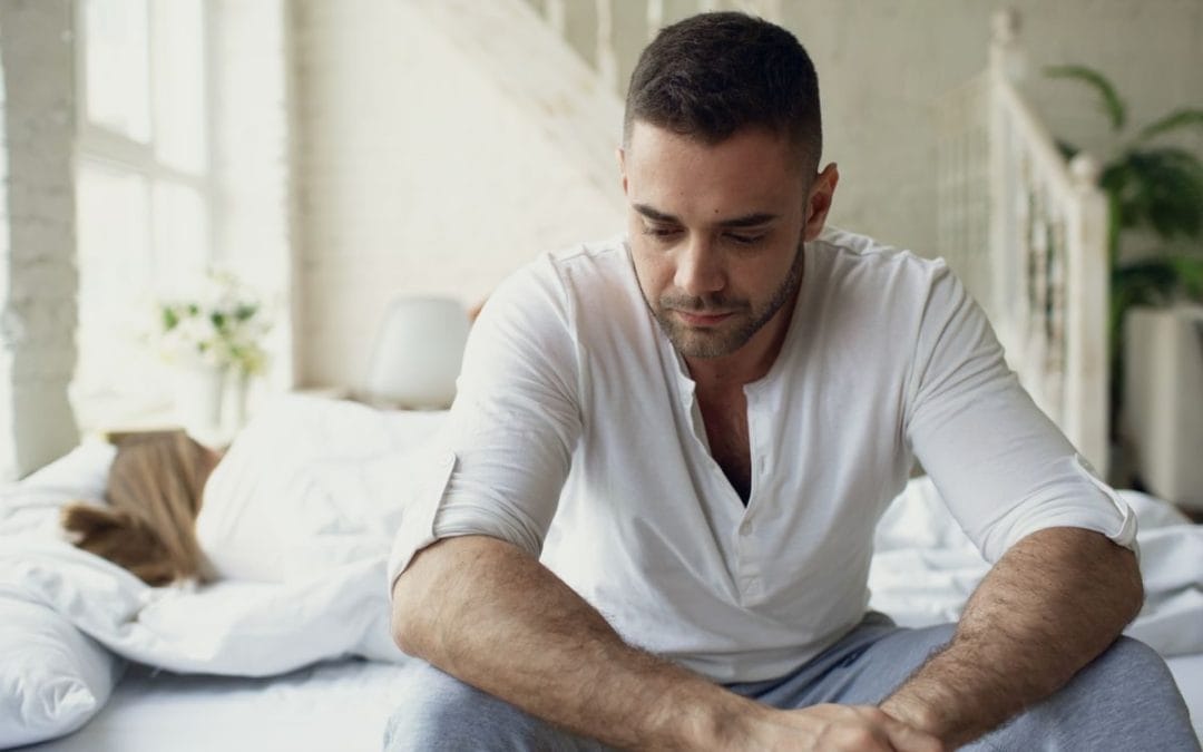 Man sitting on the edge of his bed in morning light, looking down, taking a moment before the day